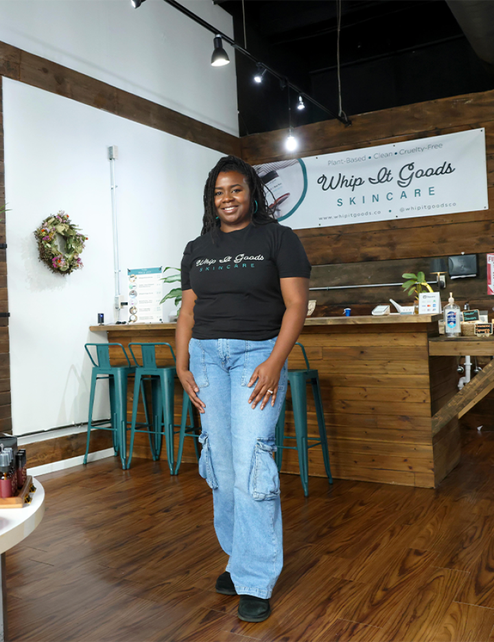 A person standing inside a store with wooden floors and walls. The person is wearing a black t-shirt with the text Whip It Goods Skincare and light blue cargo pants. Behind them is a counter with teal stools and various items on display. A sign on the wall reads Whip It Goods Skincare Specialists. There is also a wreath on the wall to the left.